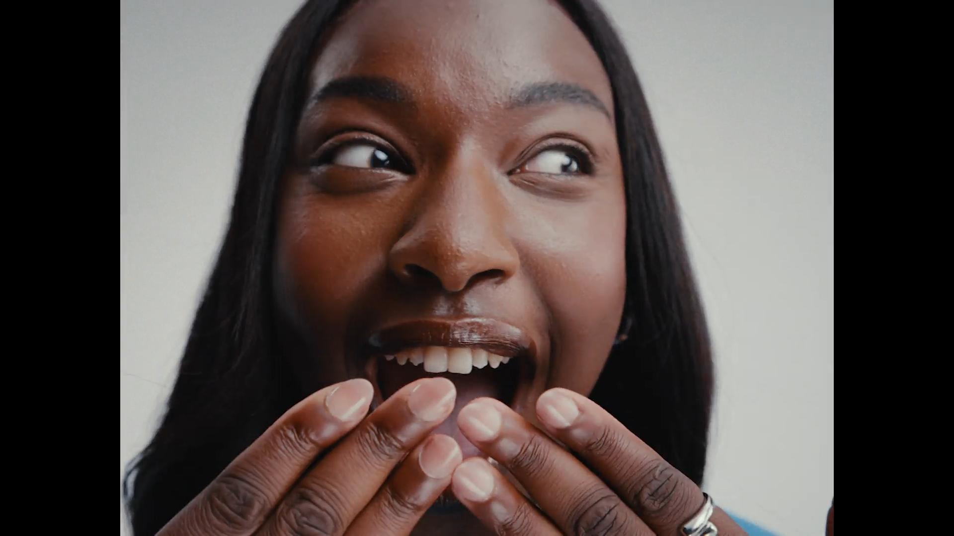Close-up of Black model with hands at mouth, eyes wide and smiling in delight.