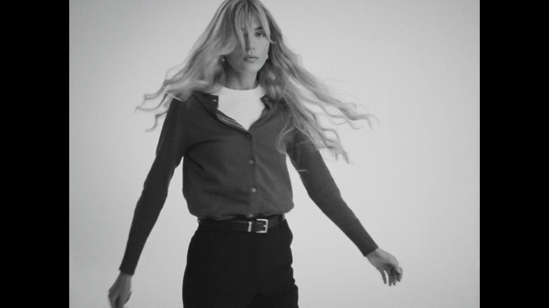 B&W of blonde model mid hair toss; dark cardigan over white tee with belt against pale cyc.
