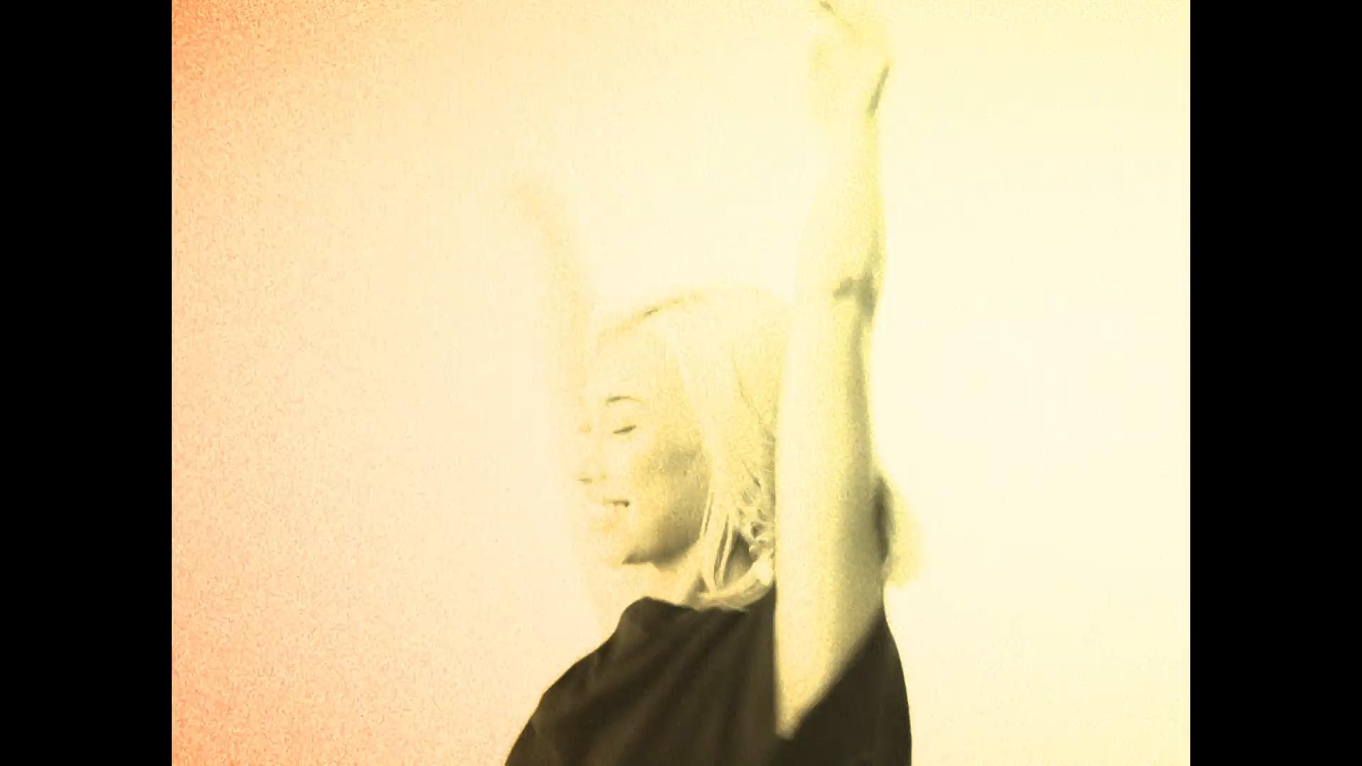 Red-tinted grainy frame of blonde model dancing with arms up and hair flying.