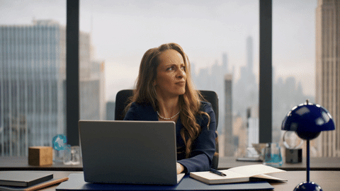 Mid-shot of the female lawyer at her desk giving a wry knowing reaction with a cobalt Verner Panton lamp right.