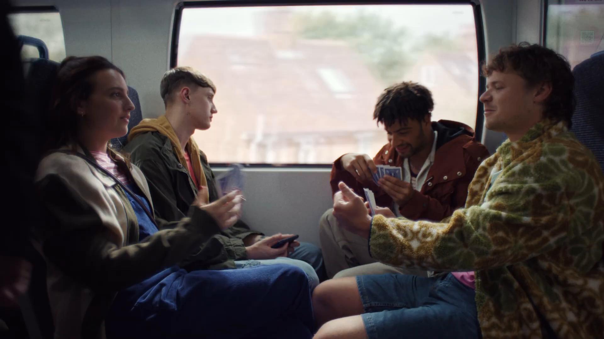 Friends playing cards on the Thameslink train — Lily, Luke, Billy and Callum at a table seat, soft countryside through the window