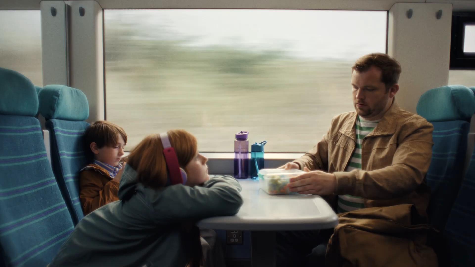 Family three-shot at a Southern train table seat — Lucas window-side, Sacha resting her head with pink-banded headphones, Dad opposite passing a packed lunch