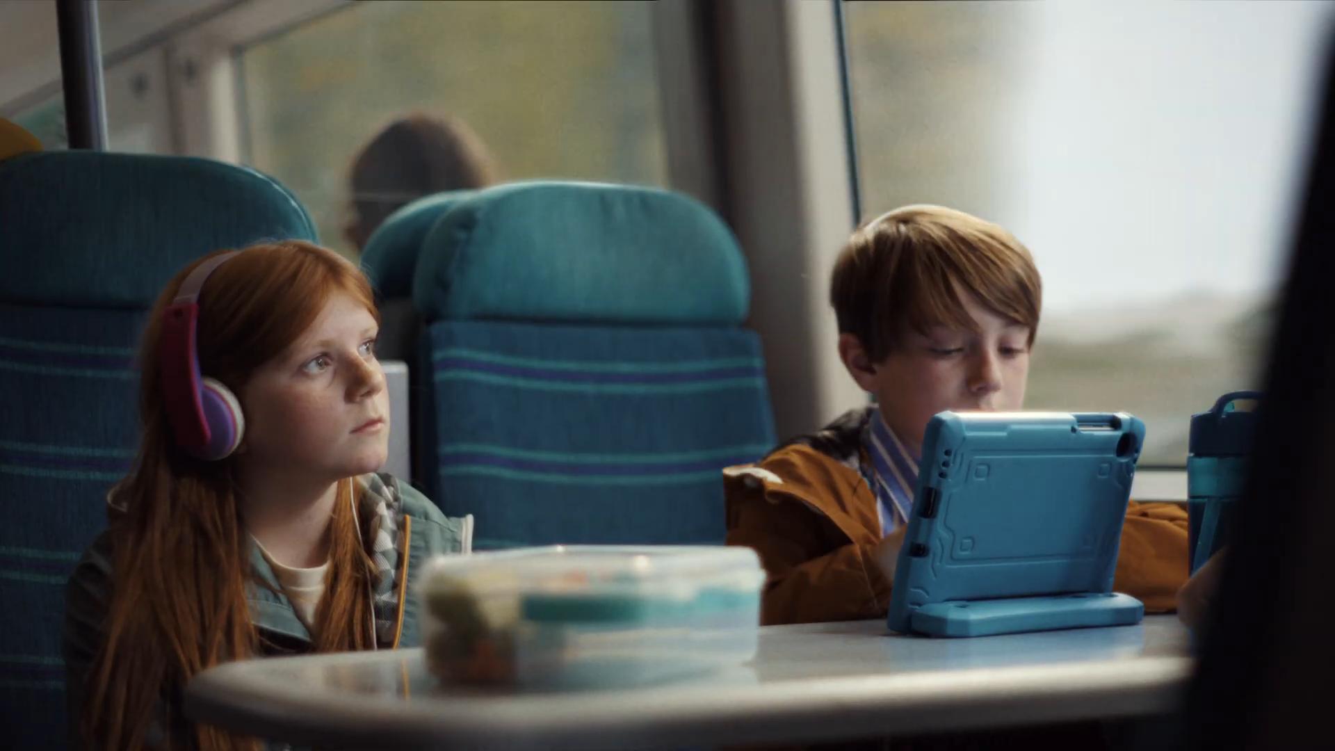 Wide of Sacha at the train table seat in headphones, Lucas opposite engrossed in his blue-cased tablet, snack box and water bottle on the table