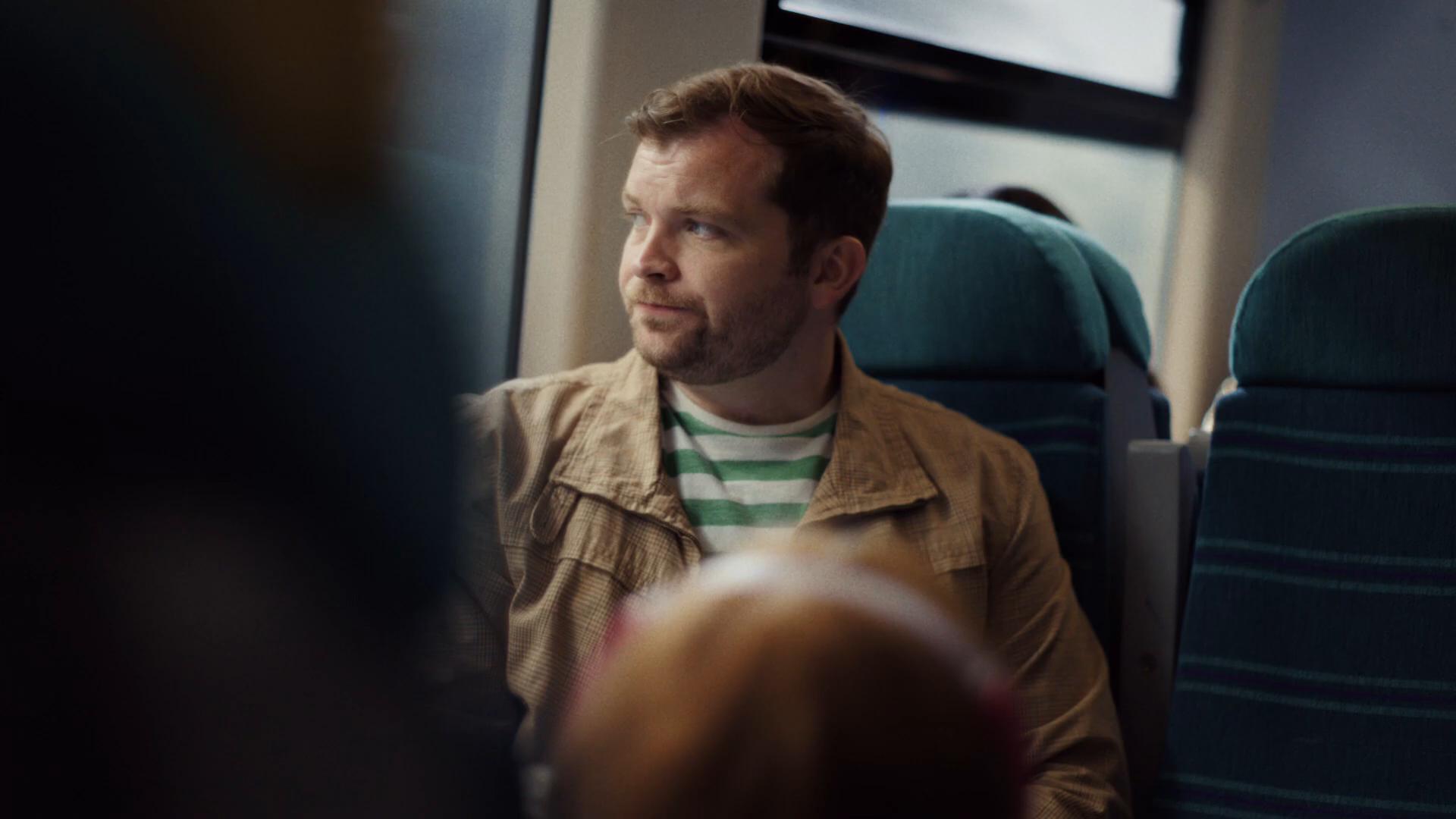Cinematic MCU of Dad on the Southern train, profile-three-quarter, looking up the carriage past the camera, the panoramic train window glowing with daylight behind him