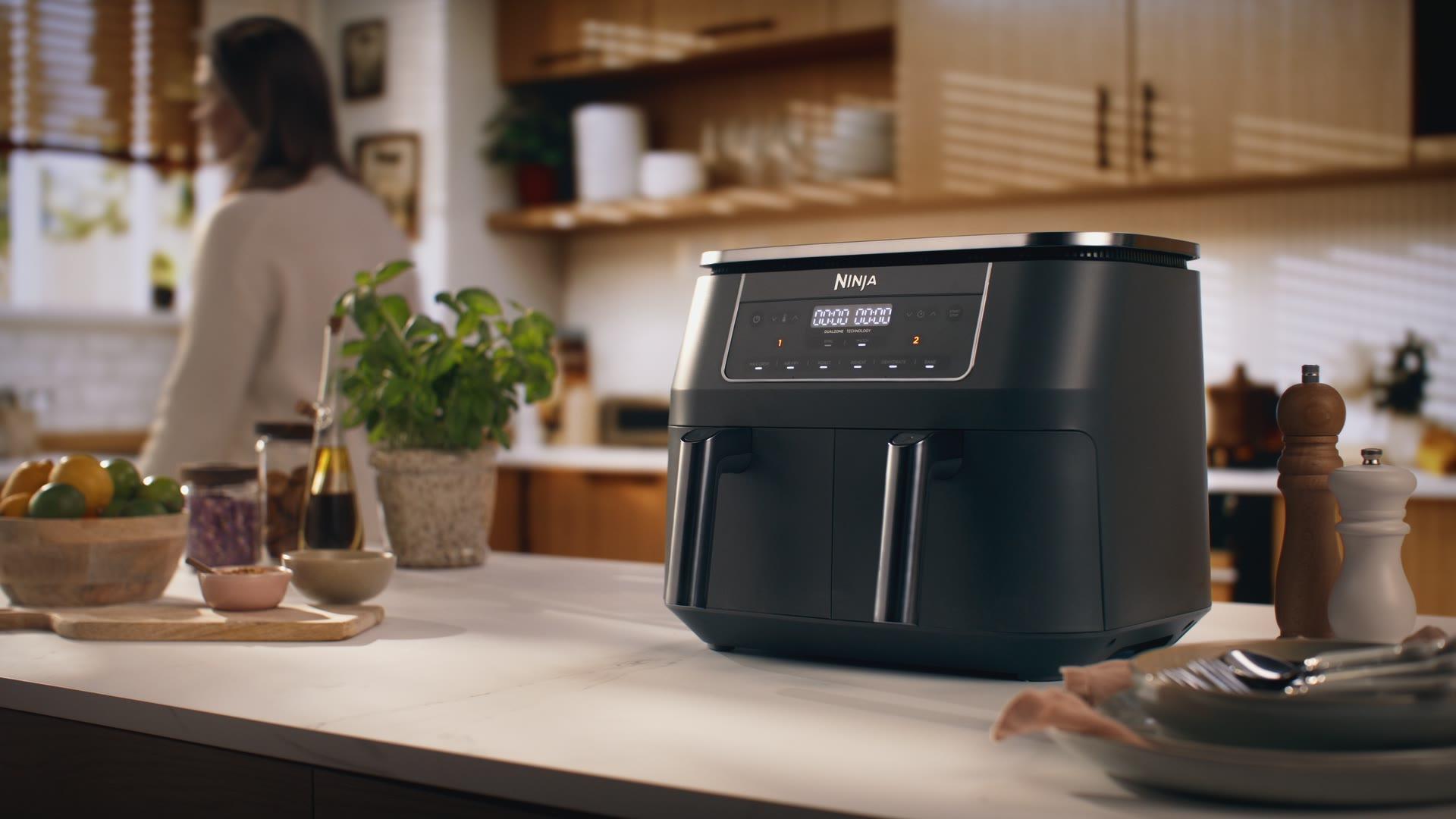 Ninja DZ300 dual-zone air fryer on a kitchen island with a woman in a cream sweater out of focus behind, lifestyle wide.