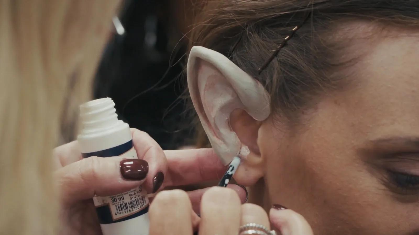 Makeup artist fits a prosthetic pointed elf ear onto a performer.